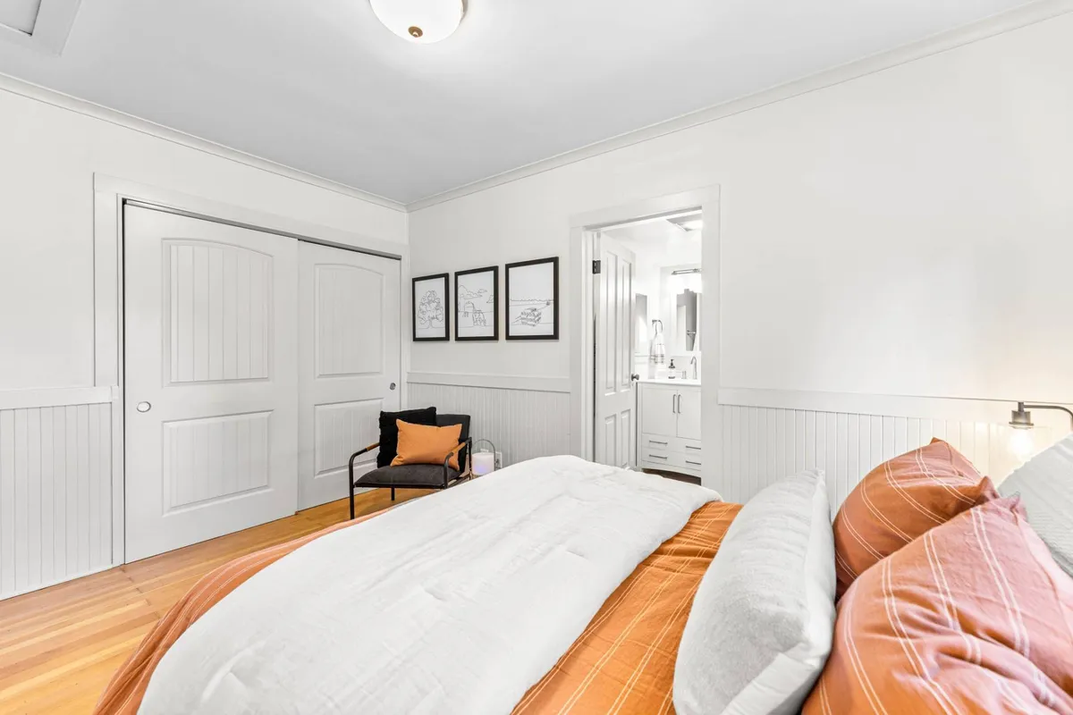 A cozy bedroom featuring a king-sized bed with orange and white bedding, complemented by soft linens and framed artwork on the wall. A chair with an orange pillow adds a pop of color, and the room is bathed in natural light, emphasizing its tranquil and restful atmosphere.