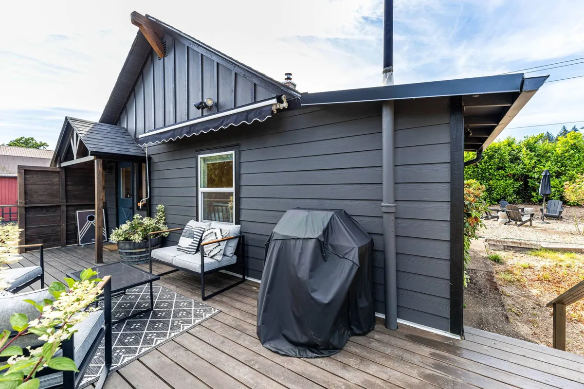 The image shows a wooden deck with outdoor seating and a grill, framed by greenery. It suggests a casual, inviting space for barbecues, starlit conversations, or enjoying a morning coffee in fresh air. The warm lighting and open layout encourage outdoor gatherings just steps from the cottage.