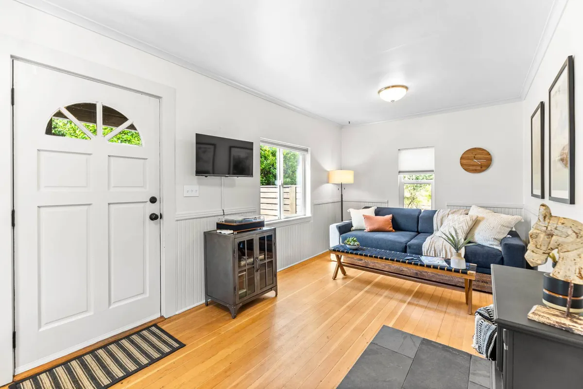 A cozy living room interior featuring a comfortable sofa, coffee table, and windows inviting natural light. The front door is visible, suggesting easy indoor-outdoor flow, while the setup offers a welcoming spot for relaxation or social gatherings.