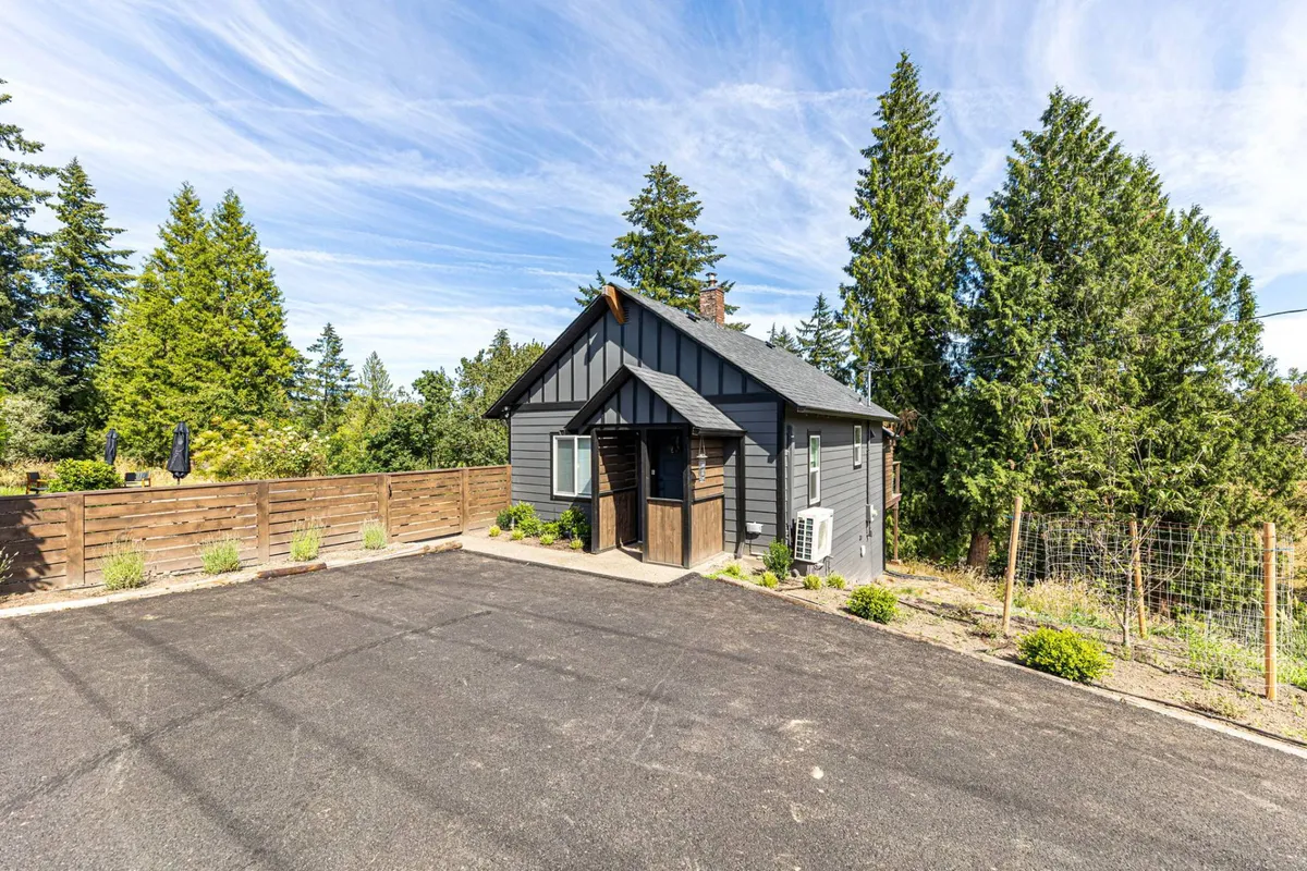 A broad view of the cottage's black and gray exterior nestled among trees and a landscaped yard, showcasing the peaceful, natural setting. The scene offers a sense of privacy and connection to the outdoors, with ample space for children to play or families to relax.