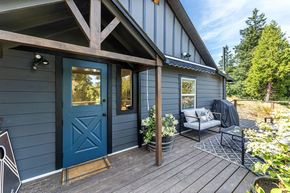 This image shows the blue front door of the cottage, an inviting outdoor seating area with cozy planters full of flowers, and a tranquil porch setting surrounded by greenery. The arrangement is perfect for relaxing, socializing, or simply enjoying the natural surroundings.