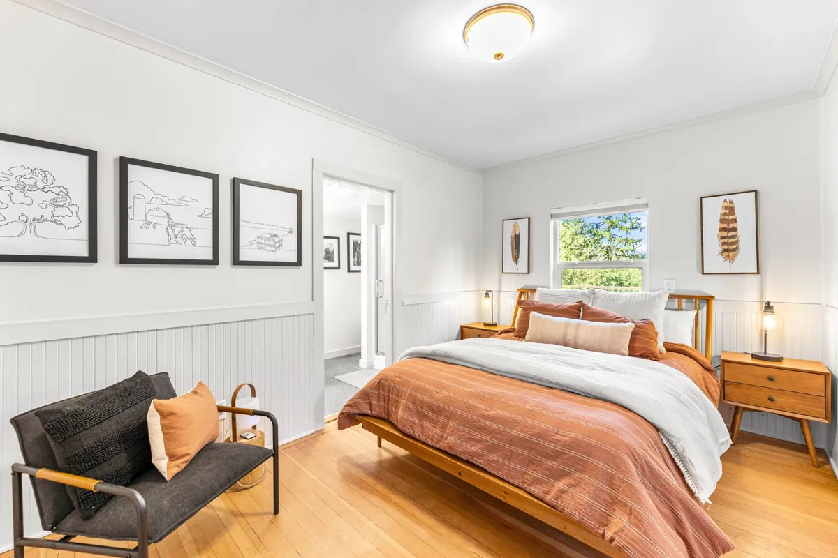 A tranquil bedroom scene with a neatly made bed featuring striped bedding, warm wood furniture, and framed artwork. Gentle natural light filters through the window, creating a peaceful atmosphere ideal for restful escapes.