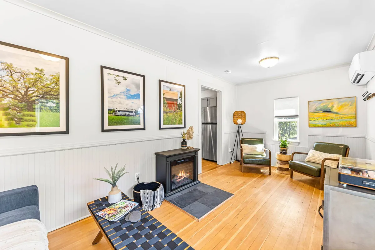 The interior shows a cozy seating area centered around a crackling fireplace, with tasteful local artwork adorning the walls. Plush chairs invite relaxation, and a small dining space suggests intimate meals, all within a warmly lit, tranquil setting.