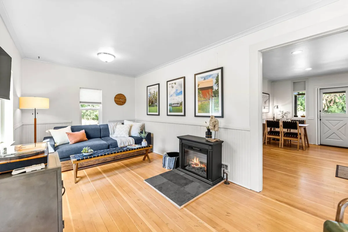 A cozy living room with a comfortable sofa, a welcoming fireplace, stylish wall art, and an inviting dining area visible in the background. The space is bathed in soft light, highlighting the new furnishings and creating a warm, communal atmosphere.